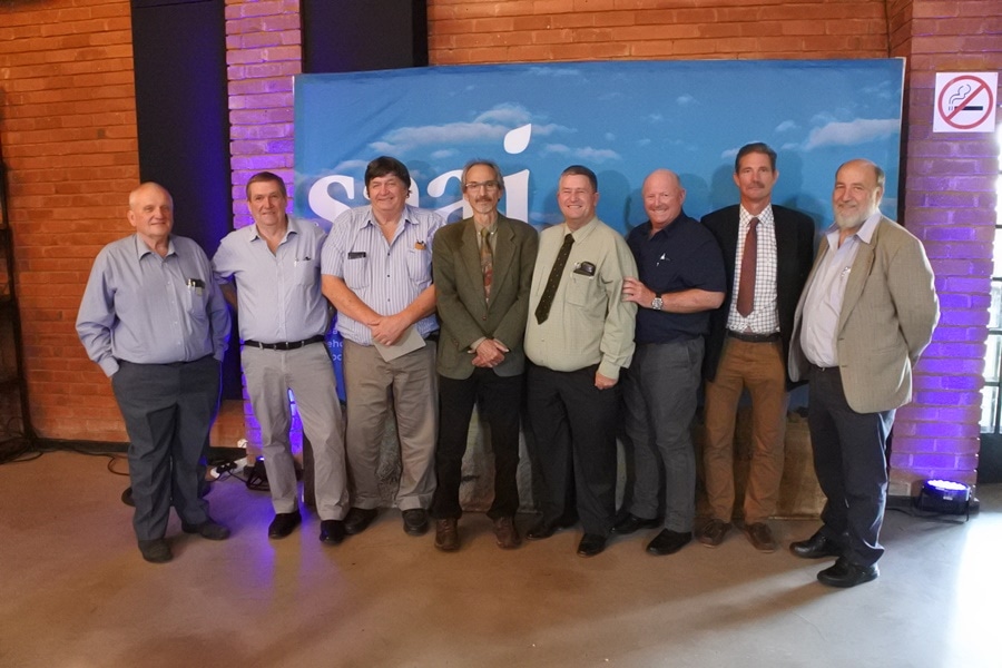Group of eight men standing side by side in business casual and formal attire against a blue Saai backdrop.