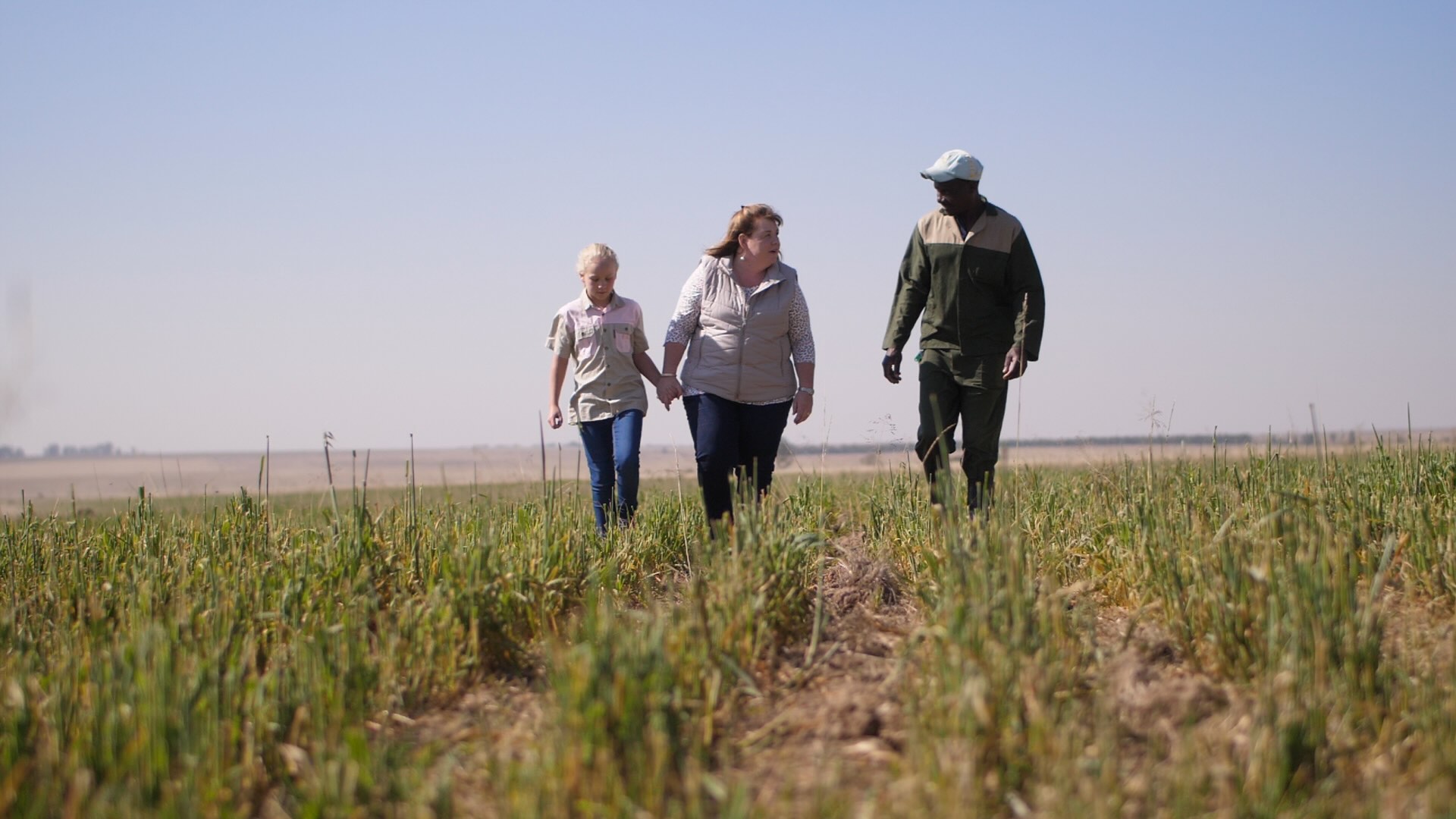 Three people walking through a green field under a clear blue sky, holding hands and engaged in conversation.