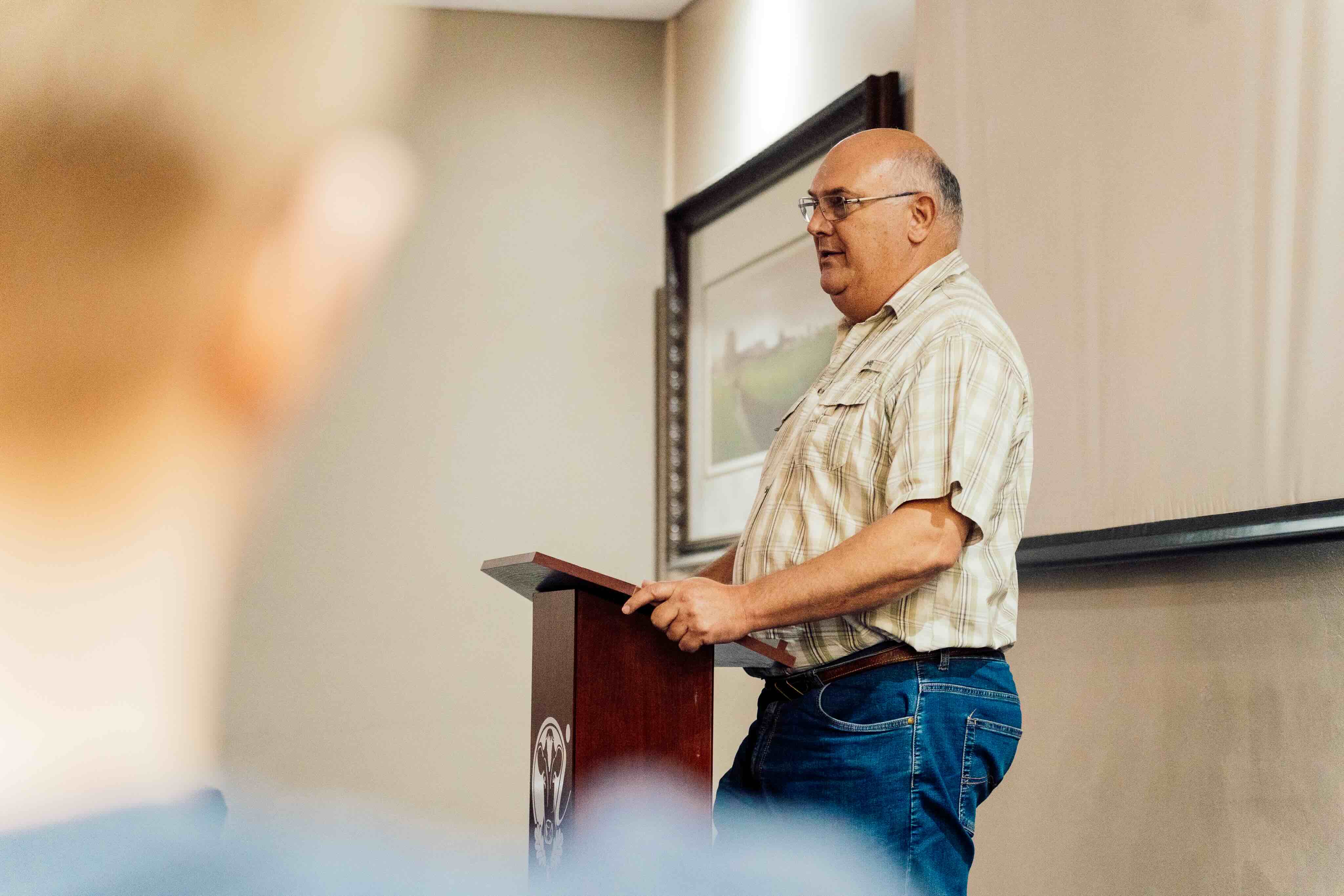Middle-aged man wearing glasses and a plaid shirt speaking at a wooden podium in a room with beige walls.