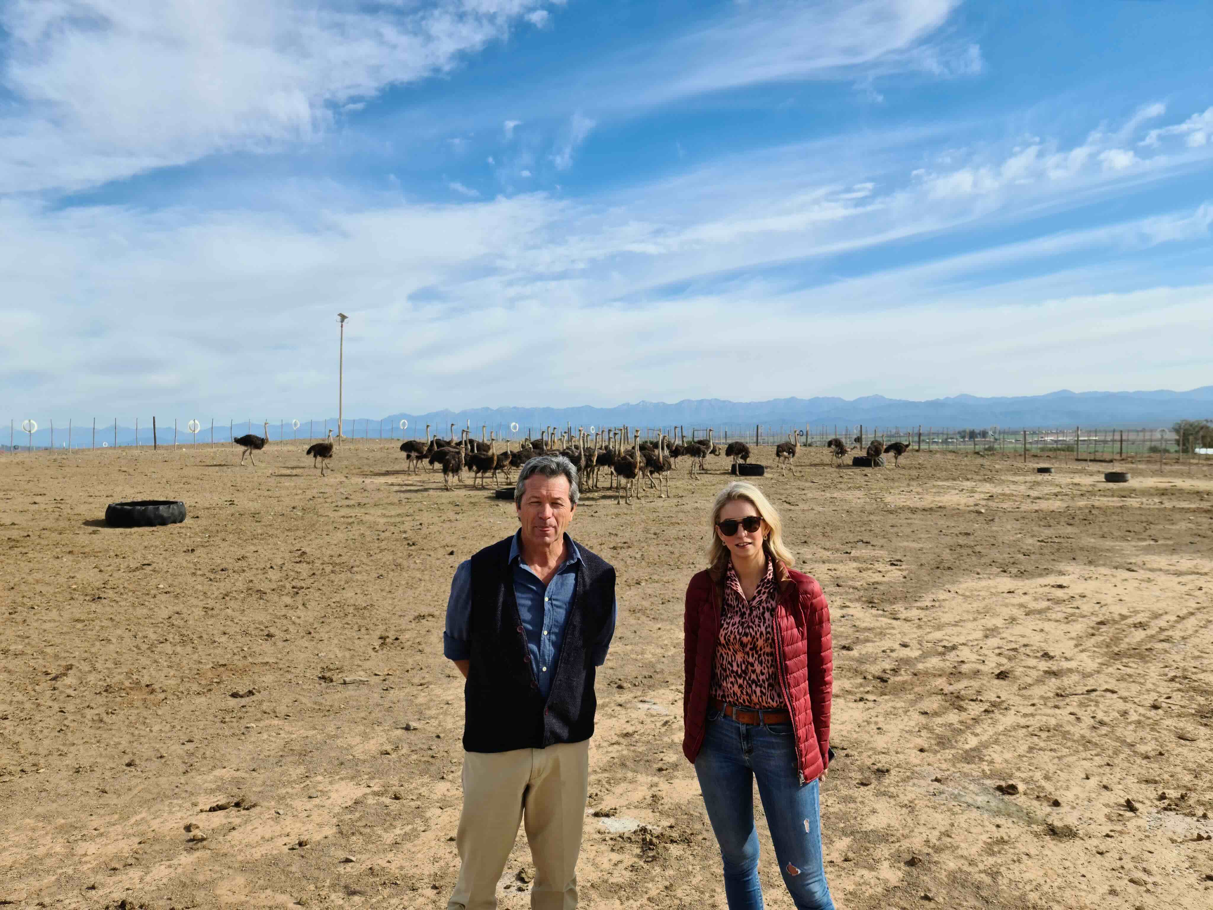Two people standing in a dry, open field with a group of ostriches in the background under a partly cloudy blue sky.