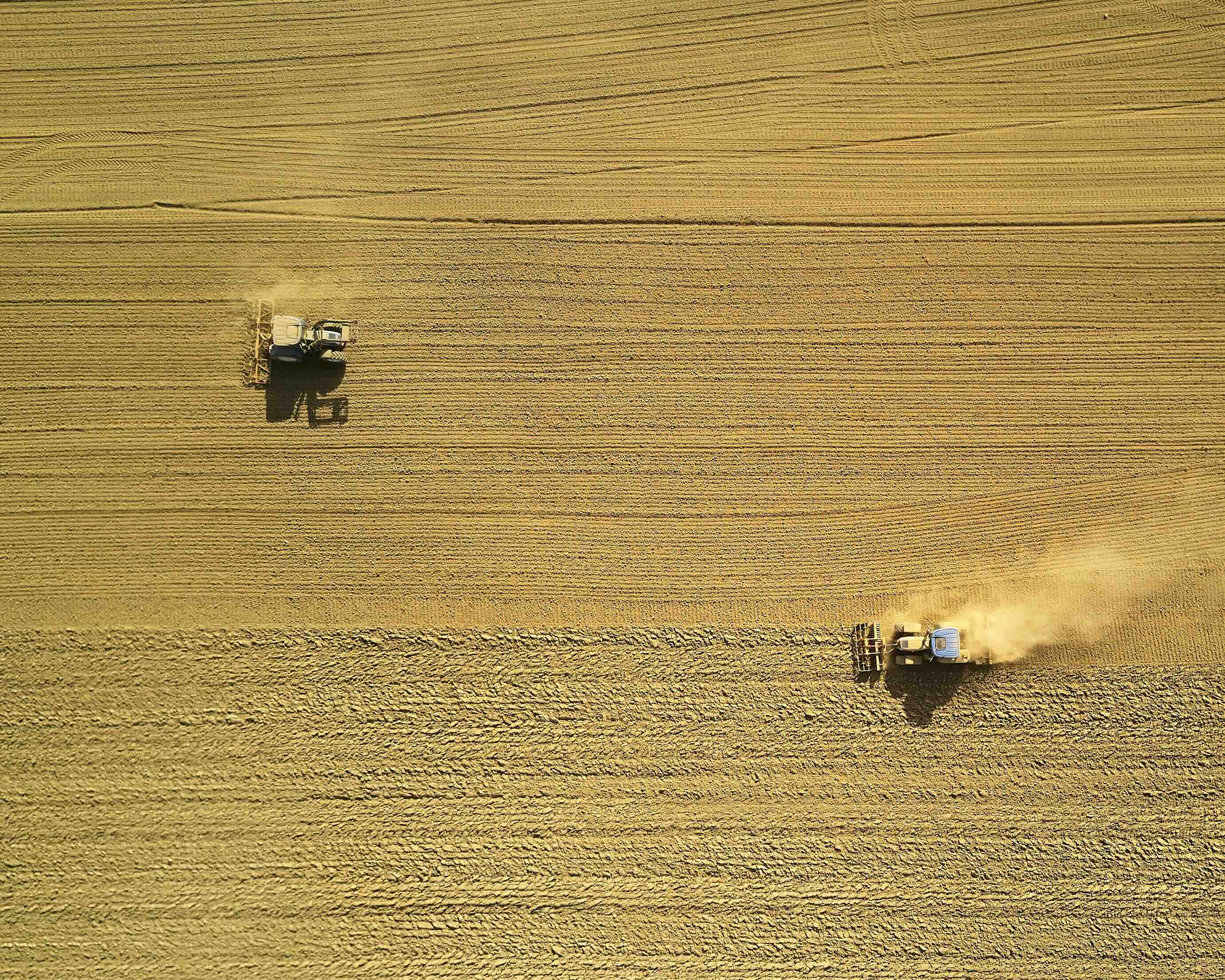 Aerial view of two tractors plowing a large golden wheat field, creating parallel lines in the soil.