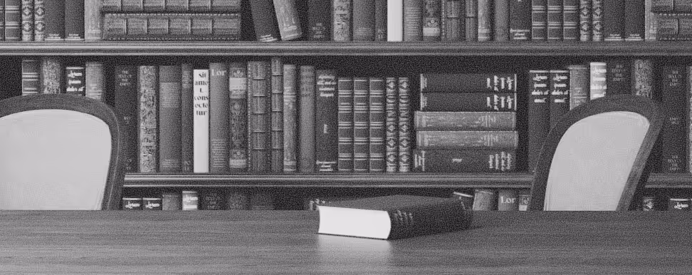 Wooden table with a closed book in front of a bookshelf filled with old books and two upholstered chairs on either side.