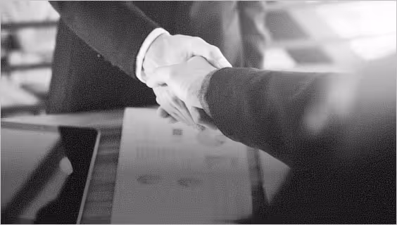 Two people in business attire shaking hands over a table with documents.