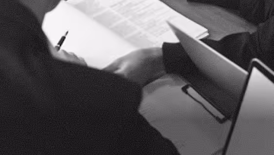 Two people reviewing documents with a pen, laptop, and clipboard on a table in black and white.