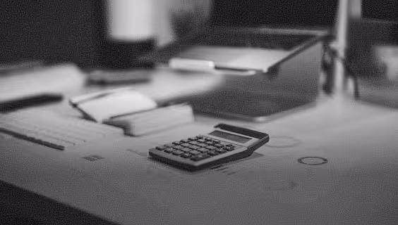Black calculator on a desk next to scattered papers with a laptop in the background.
