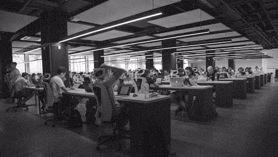 Large open office space with rows of desks where people are working on laptops and computers.
