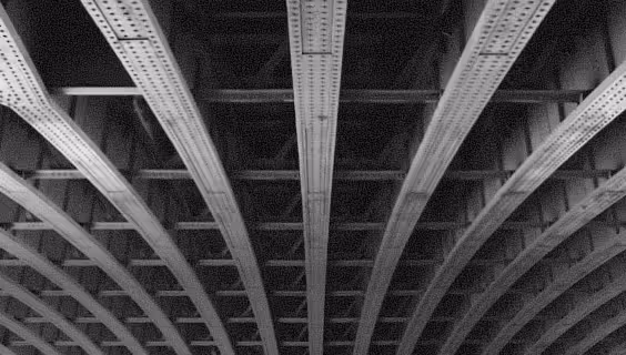 Black and white photo of metal beams and riveted girders forming the underside of a bridge or industrial structure.