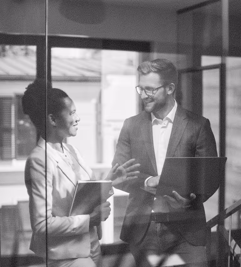 Two business professionals, a woman holding a folder and a man holding a laptop, smiling and conversing in an office setting.