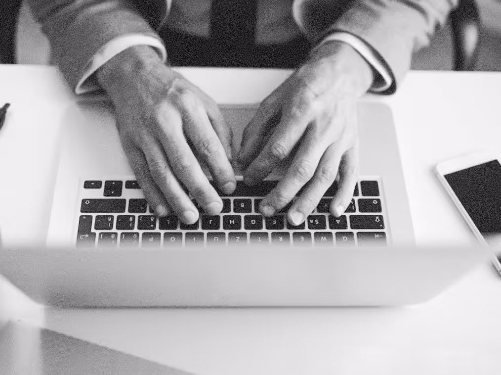 Black and white photo of a person typing on a laptop keyboard with a smartphone on the desk nearby.
