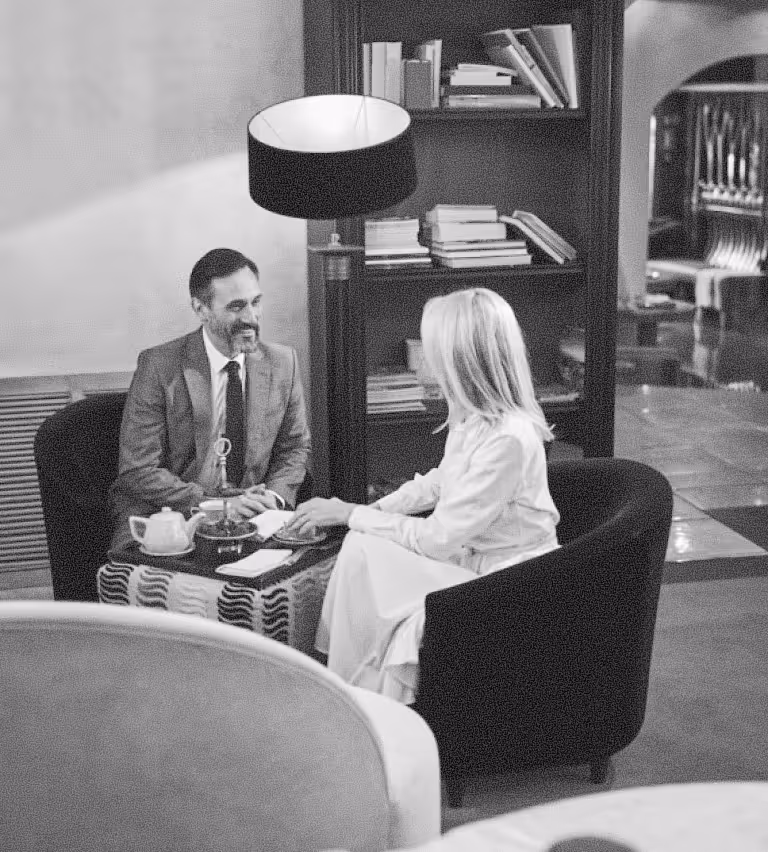 Man in a suit and woman in a dress sitting in armchairs having tea and conversation in a cozy room with bookshelves.
