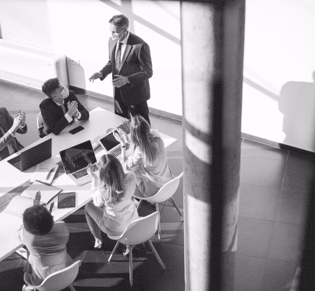 Business meeting with five people wearing masks seated around a table with laptops and documents, one man standing and speaking.