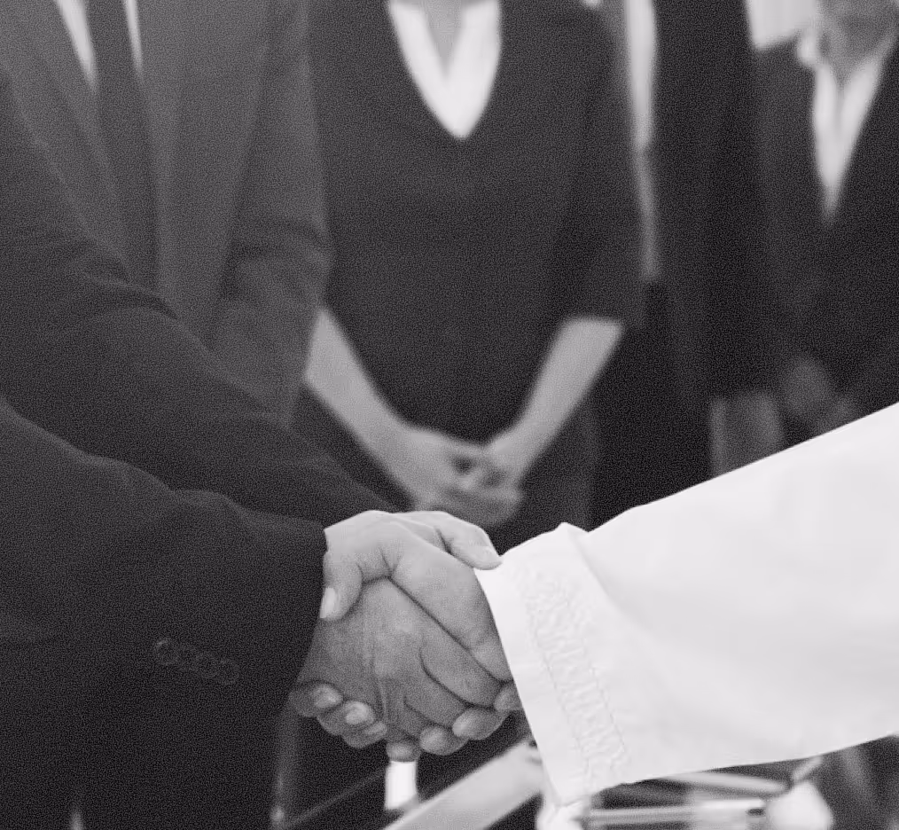 Two people shaking hands, one wearing a dark suit and the other a white traditional garment.