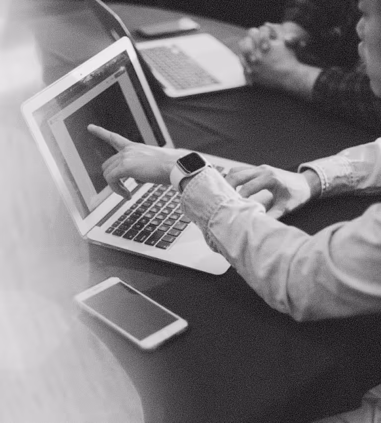 Person wearing a smartwatch pointing at a laptop screen while another person watches, with a smartphone on the table.