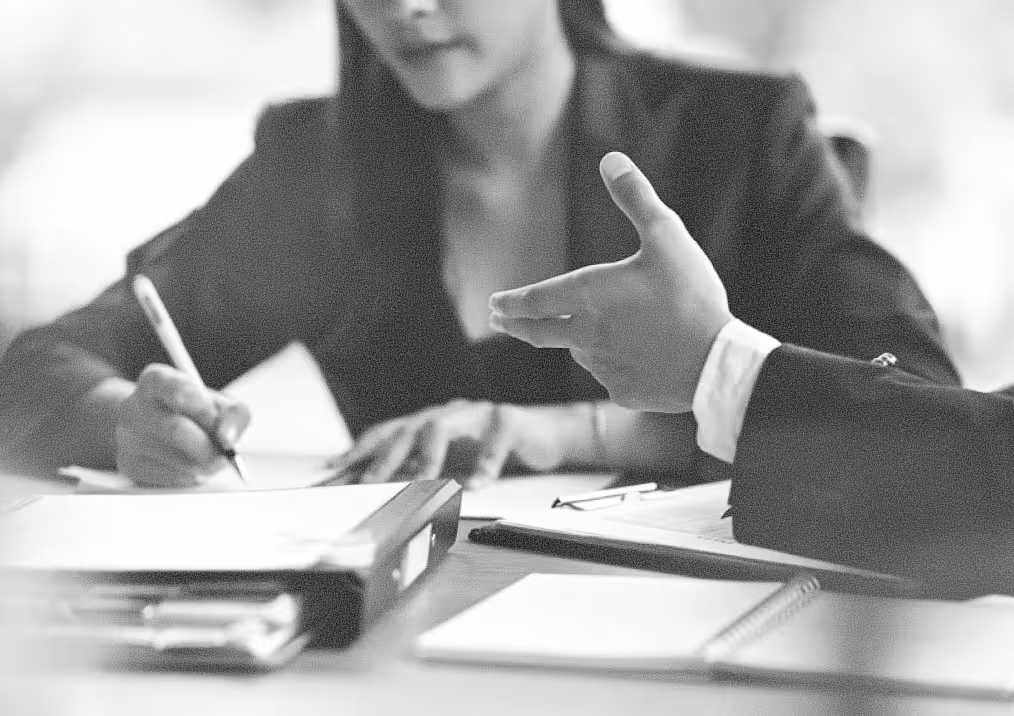 Two business professionals engaged in a discussion with documents and notebooks on the table, one person writing and the other gesturing with their hand.