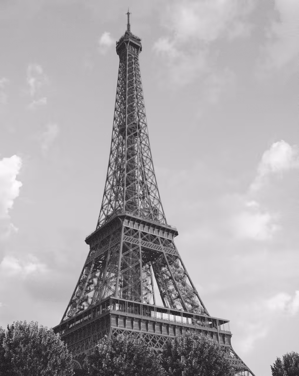 Black and white image of the Eiffel Tower rising above tree tops under a cloudy sky.
