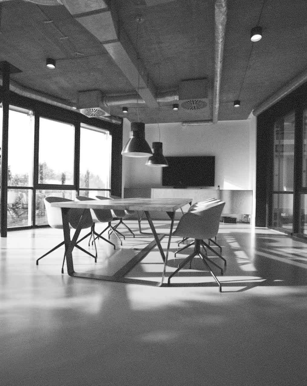Modern conference room with a long table, swivel chairs, pendant lights, and large windows casting shadows on the floor.