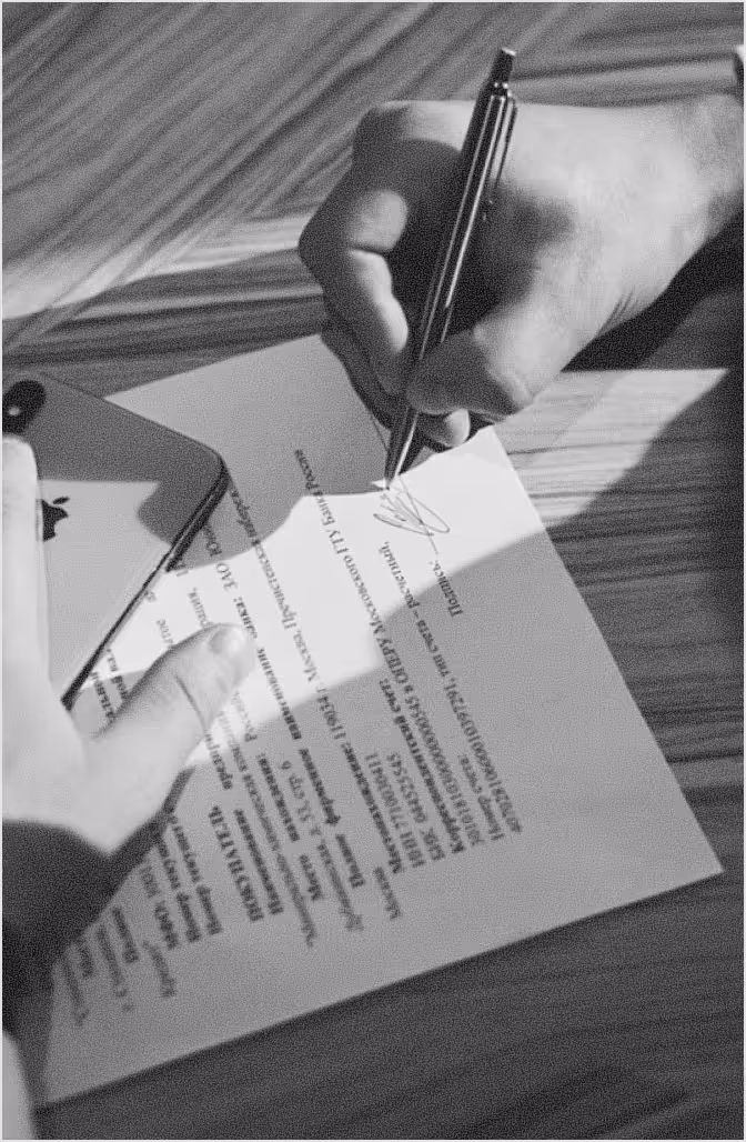 Person signing a document on a wooden surface with a smartphone nearby.