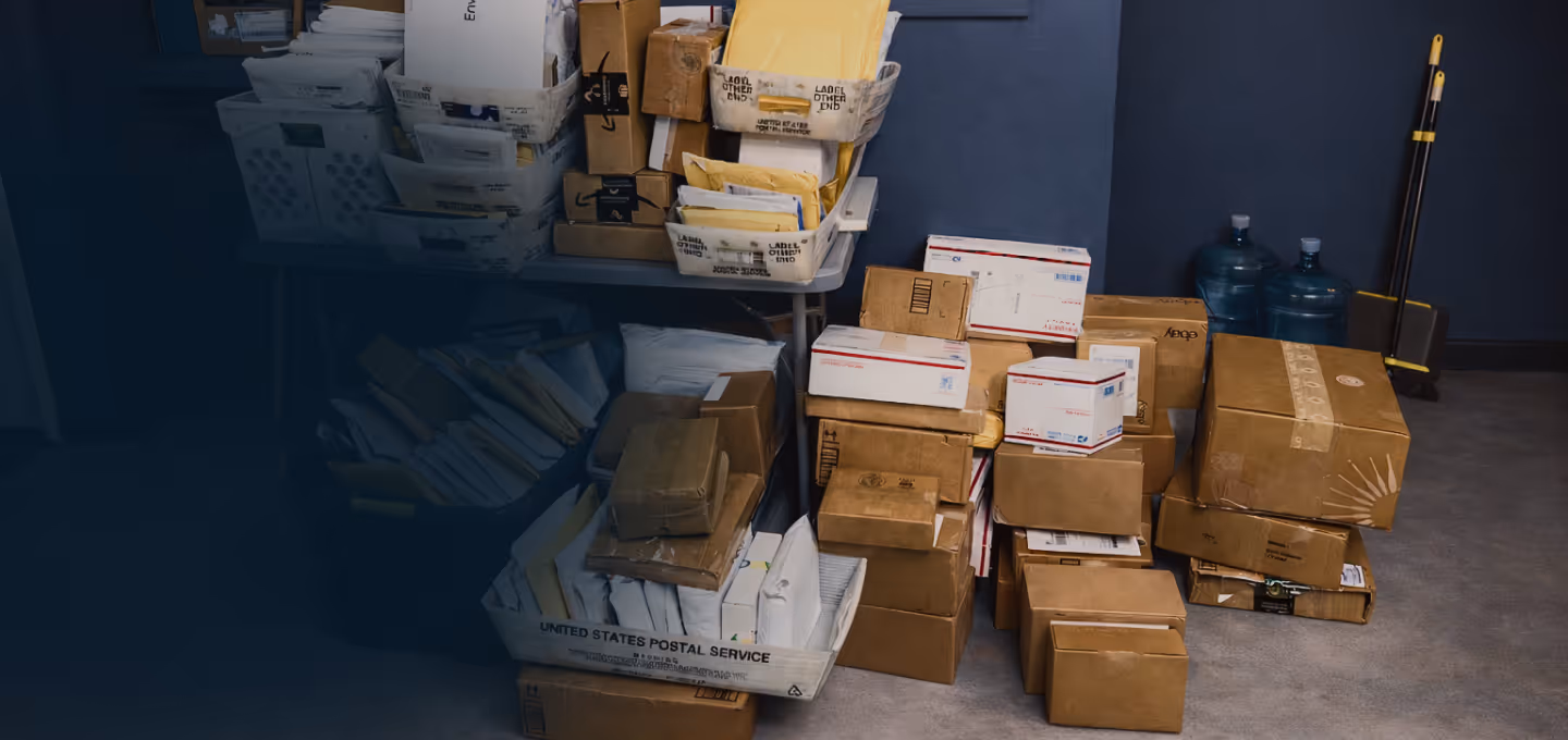 Stack of various cardboard boxes and padded envelopes in a room with two water jugs and cleaning tools in the background.