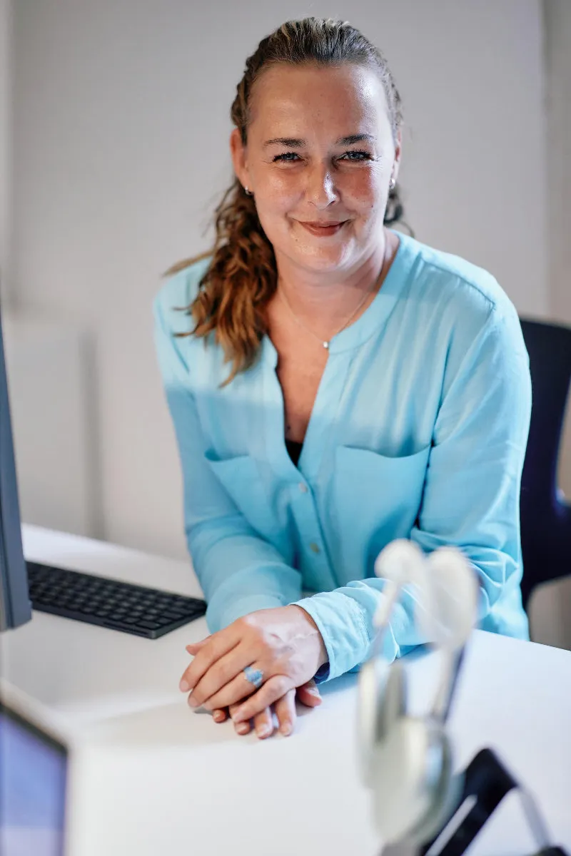 Susanne Gründerin von Pagepass mit langem, zurückgebundenem Haar und blauem Hemd lächelt am Schreibtisch im Büro.