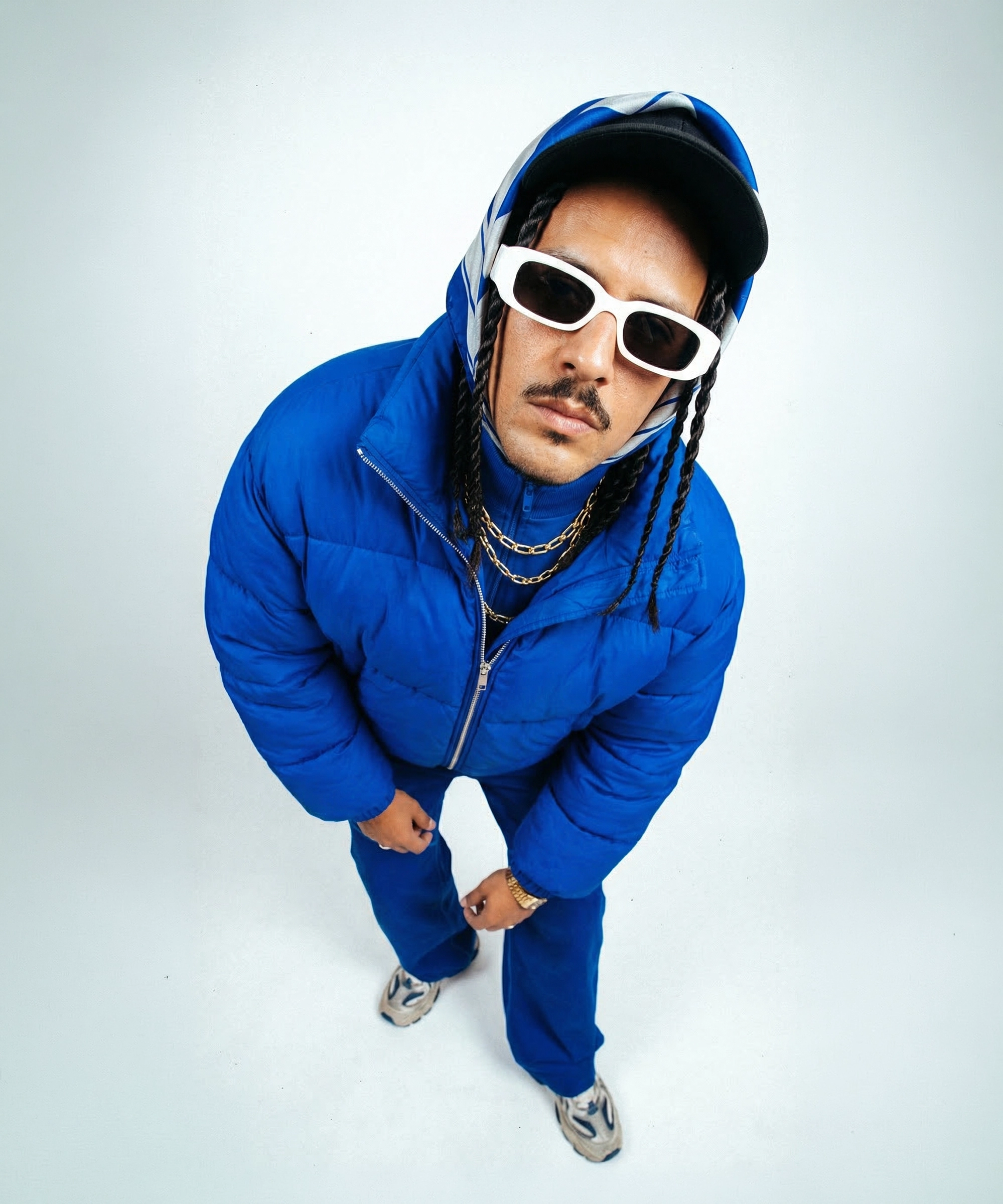 Man wearing white sunglasses, blue puffer jacket, blue pants, black cap, and a blue patterned scarf, posing against a plain white background.