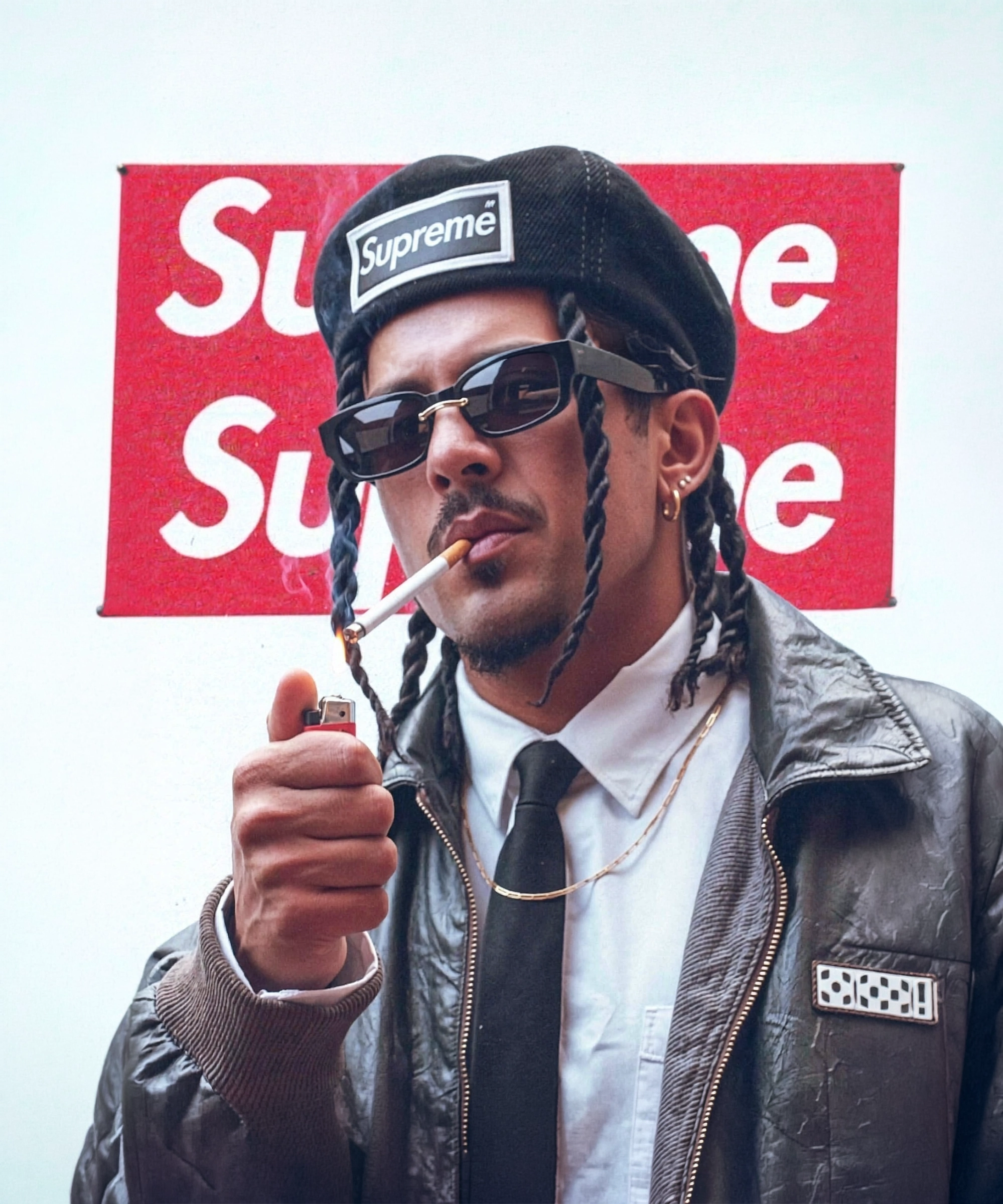 Man with braided hair wearing a Supreme hat, sunglasses, leather jacket, and smoking a cigarette while lighting it with a lighter.