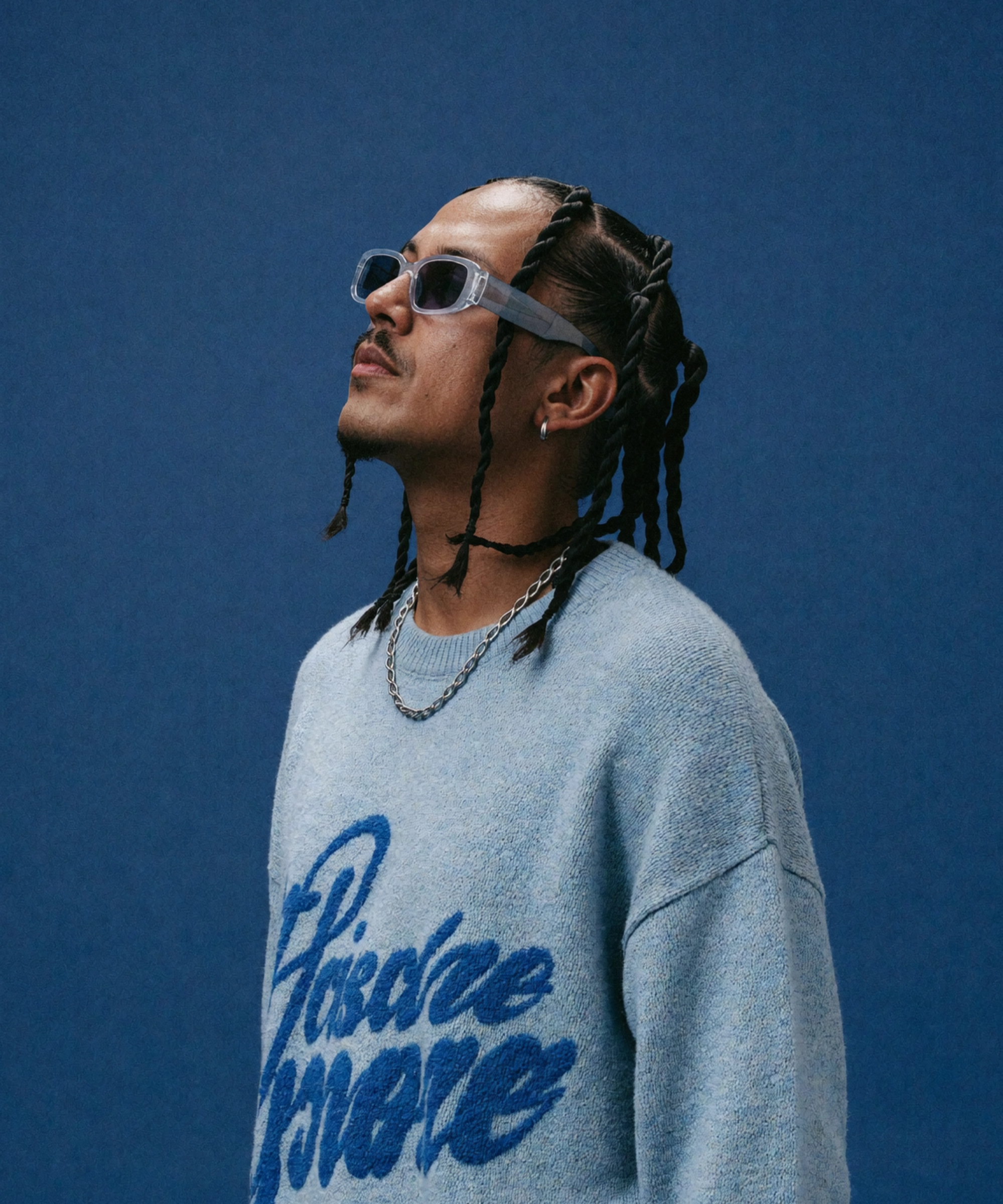 Man with braided hair and wearing rectangular sunglasses, a chain necklace, and a blue sweater with large script text looking upward against a plain blue background.