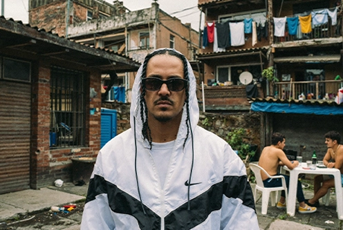 Man with sunglasses and braided hair wearing a white and black jacket standing in an urban courtyard with brick buildings and people sitting at a table in the background.