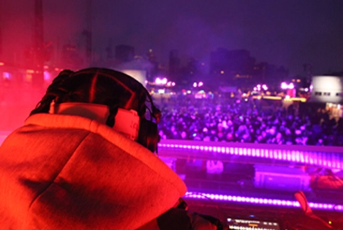 DJ wearing headphones and hoodie performing at a nighttime event with a large crowd and purple lighting in the background.