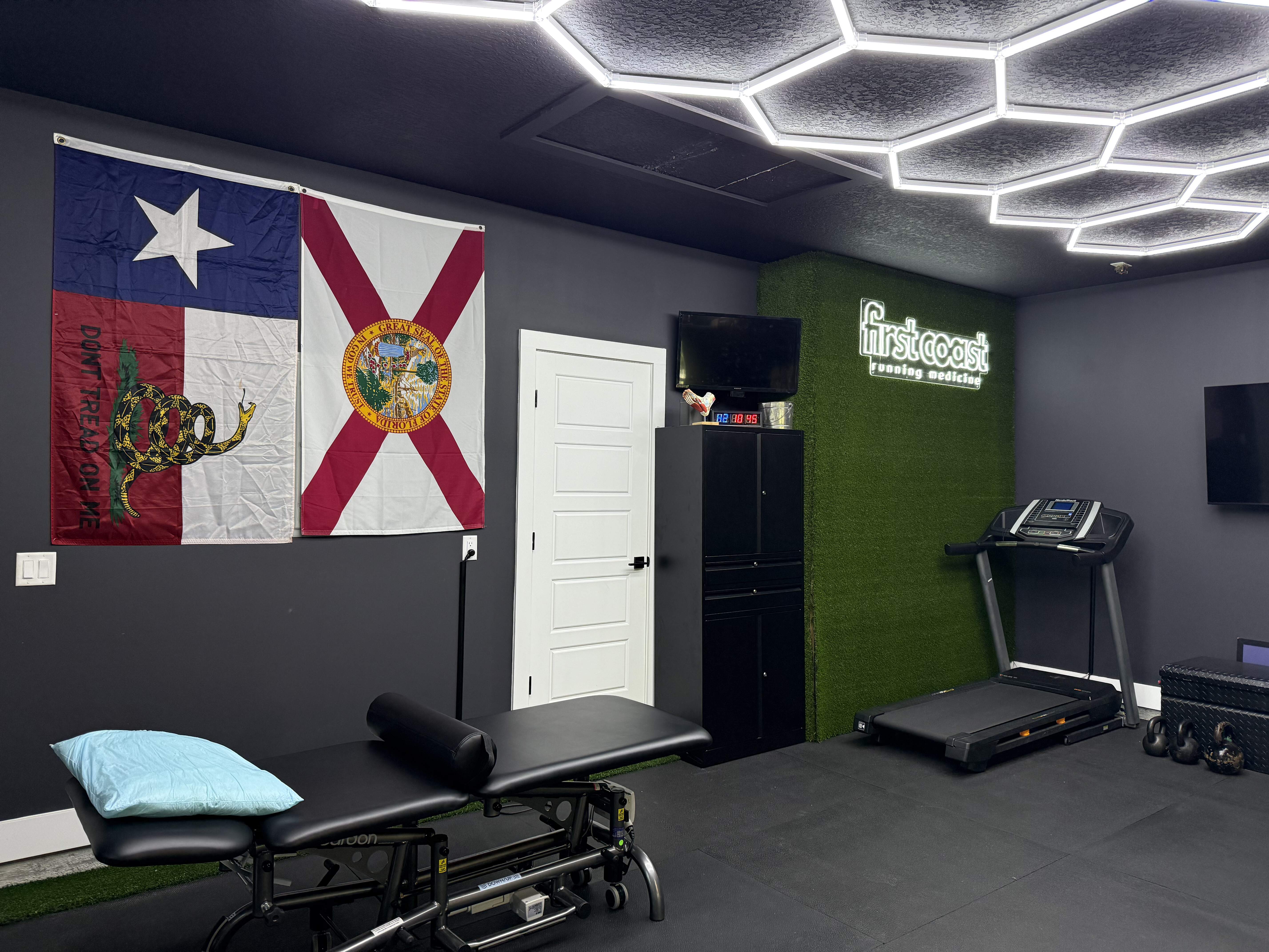 Modern physical therapy room with a black treatment table, Texas and Florida flags on wall, treadmill, kettlebells, and illuminated First Coast Running Medicine sign.