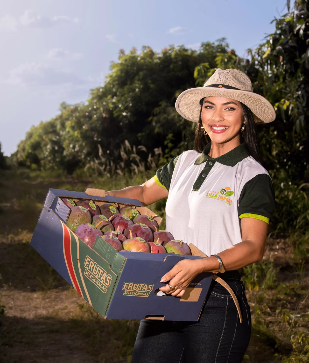 Smiling woman in hat holding a box of ripe mangoes in an orchard on a sunny day.