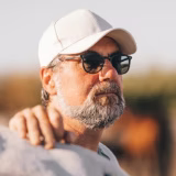 Bearded man wearing sunglasses and a white cap outdoors with blurred background.