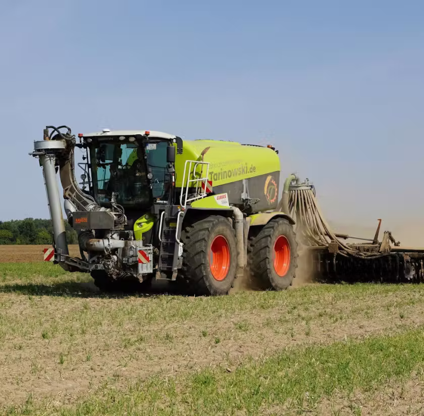 Large green Claas tractor working on a farm field under clear sky, pulling farming equipment.