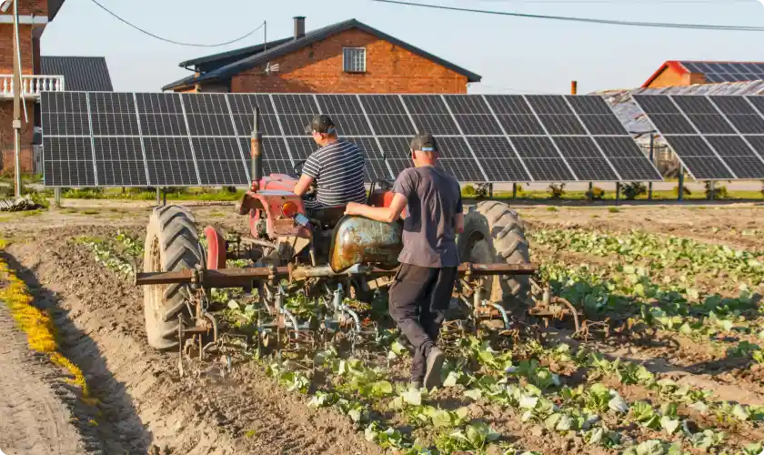 Two farmers working on tractor in a field with solar panels and houses in the background.
