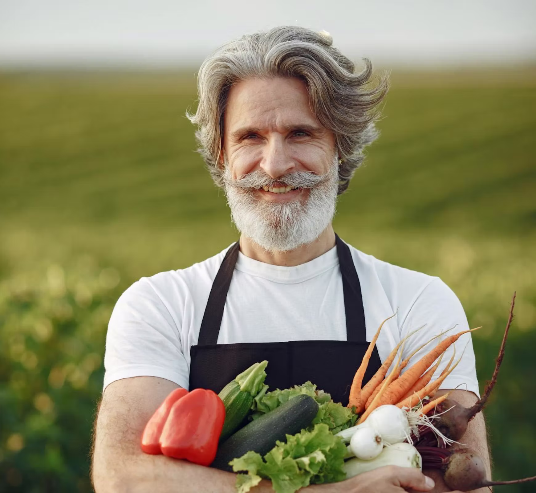 Smiling older man with gray hair and beard holding fresh vegetables in a field.