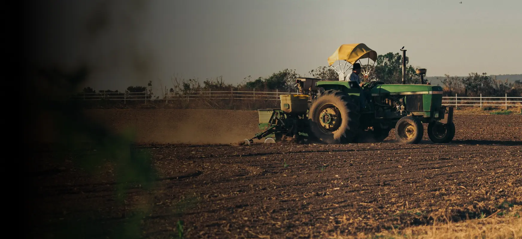 Green tractor with yellow canopy driven on dry farmland under clear sky at sunset.