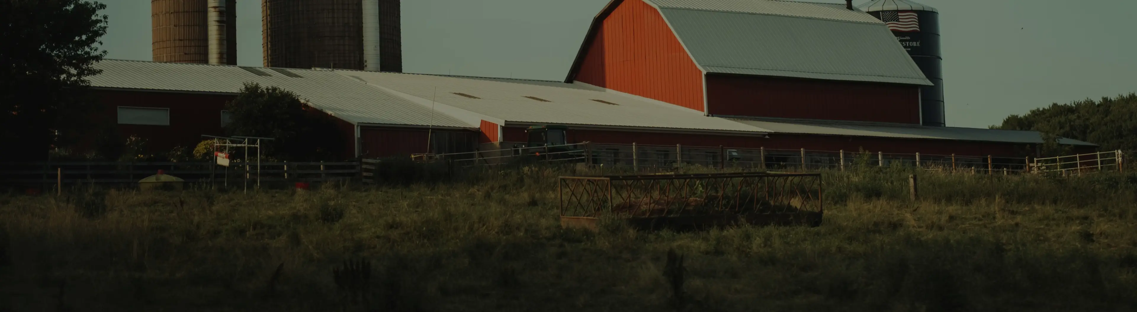 Red barn with silver roof, silos, and fenced grassy area on a farm at dusk.