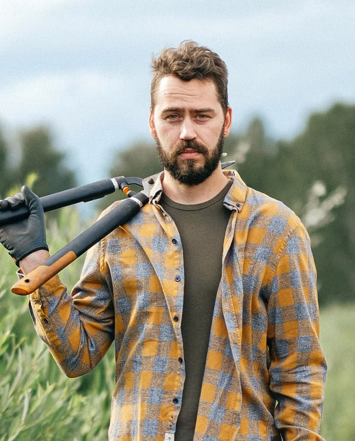 Bearded man in yellow plaid shirt holding garden shears over his shoulder outdoors.