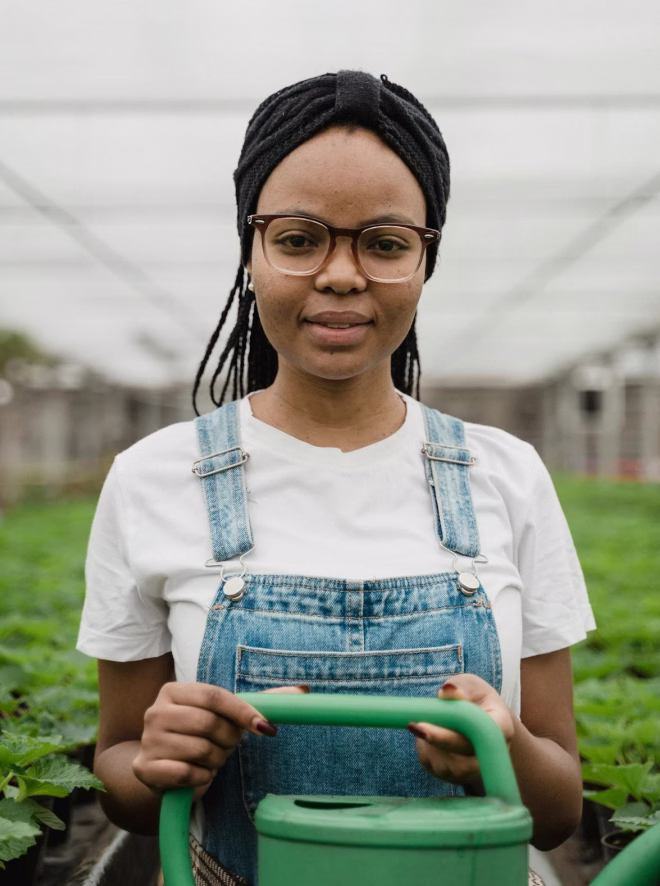 Woman in glasses and denim overalls holding a green watering can in a greenhouse.