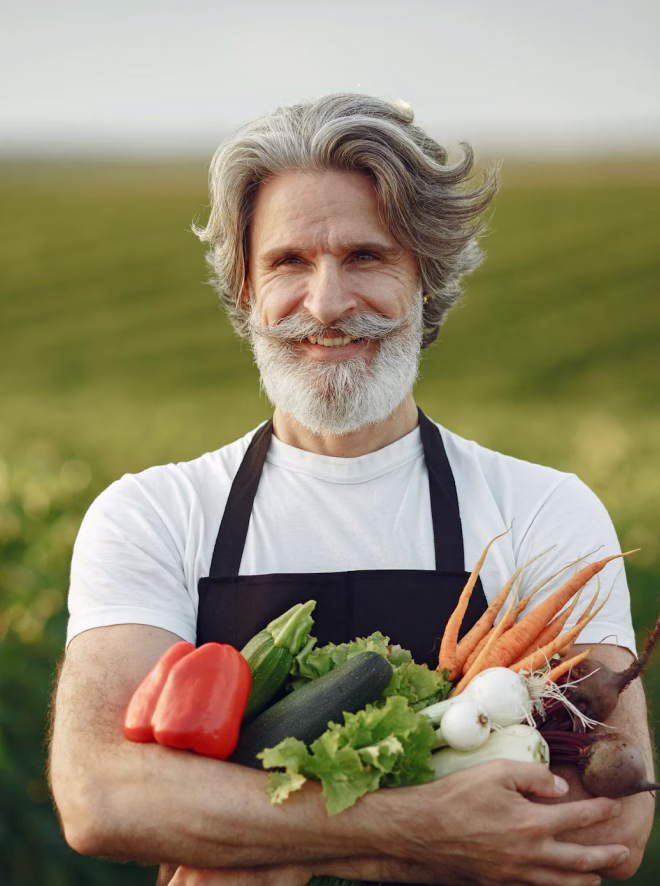 Smiling older man with white beard holding fresh vegetables outdoors in a field.
