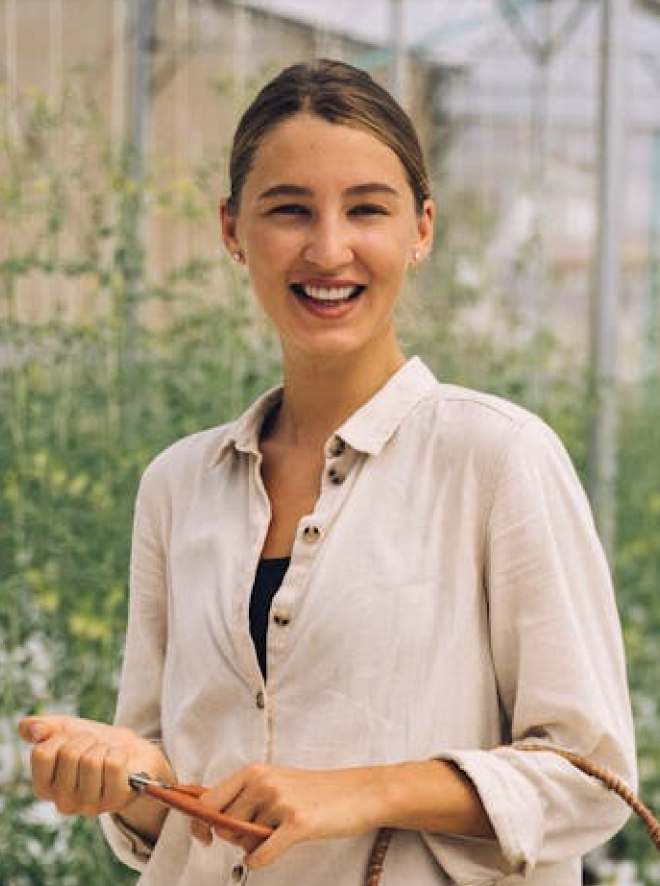 Smiling woman in beige shirt holding a pen, standing in a greenhouse with plants in the background.