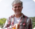Smiling older man in a plaid shirt holding a sandwich outdoors.