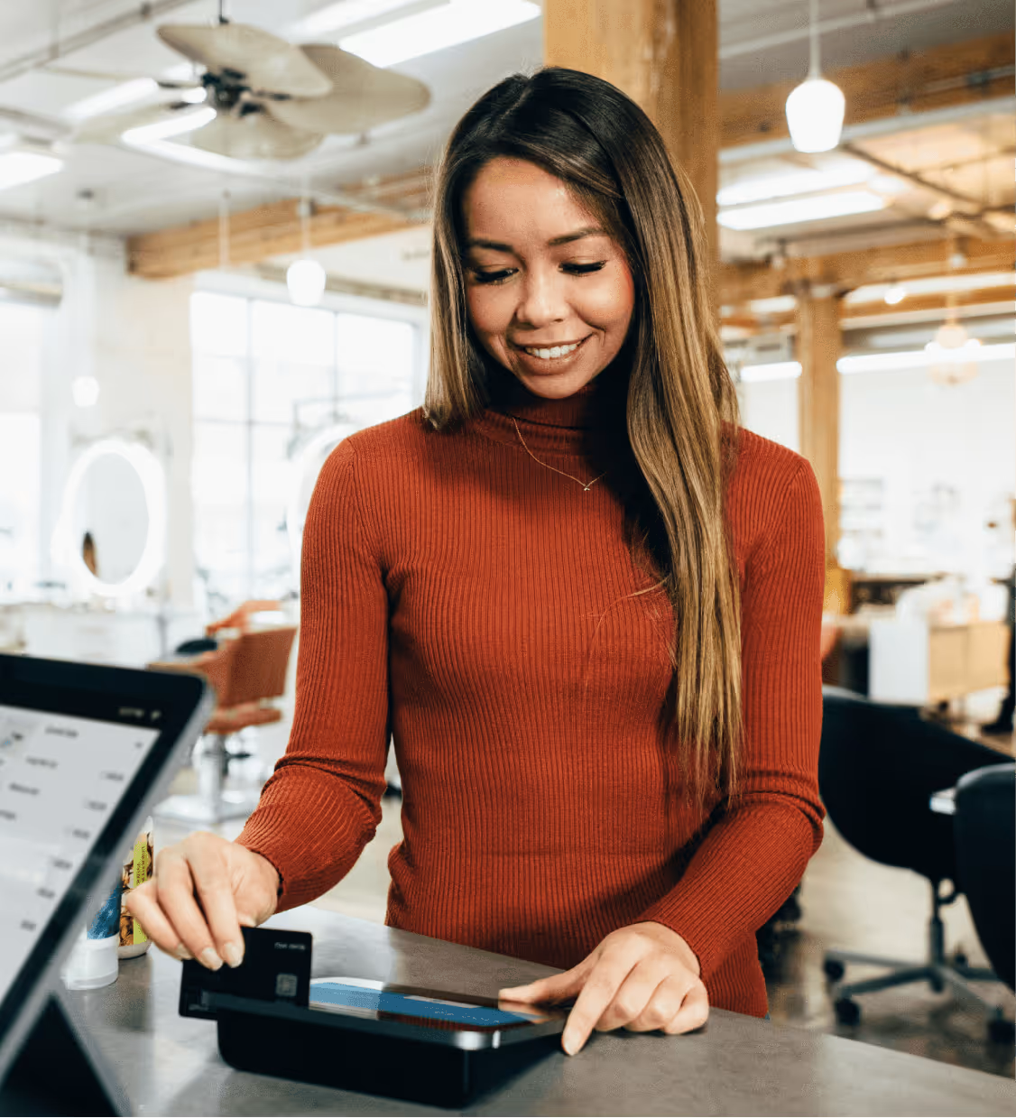 Smiling woman in red sweater making a contactless card payment using a card reader in a modern indoor setting.