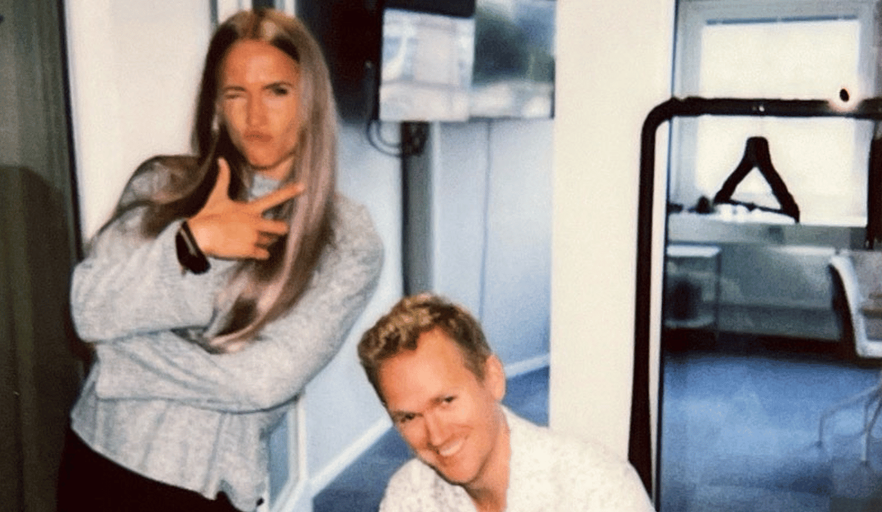 Två personer ler, en kvinna gör en fredsteckenpose inne i ett rum med soffor och en spegel.