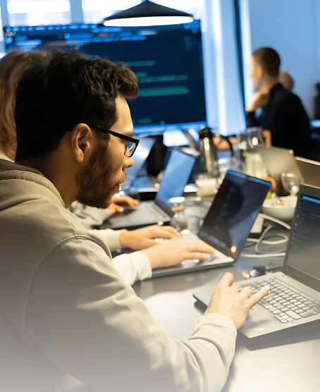 Person with glasses working on a laptop in an office with others and code on screens.
