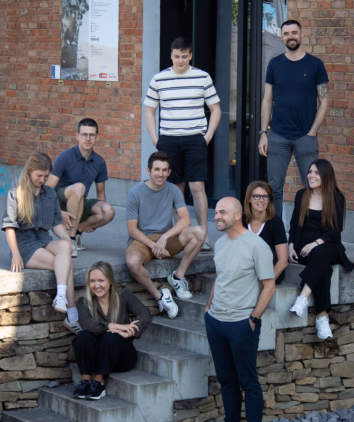 Groep van negen mensen zit en staat buiten op trappen bij een bakstenen gebouw, lachend en ontspannen.