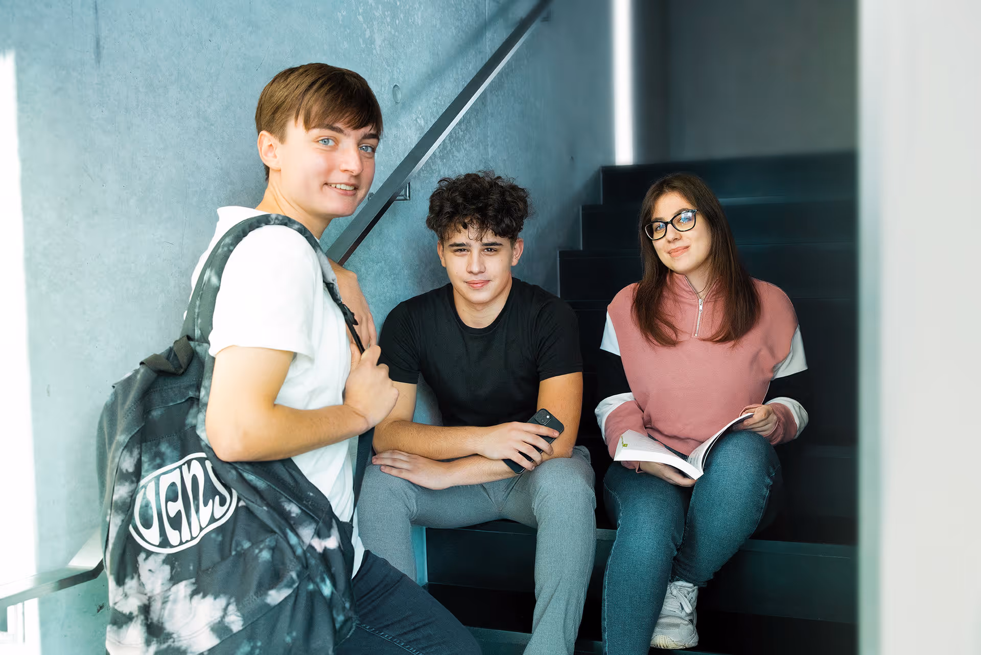 Drei junge Studenten sitzen auf der Treppe; einer mit einem Rucksack, einer mit einem Telefon und einer liest ein Buch.