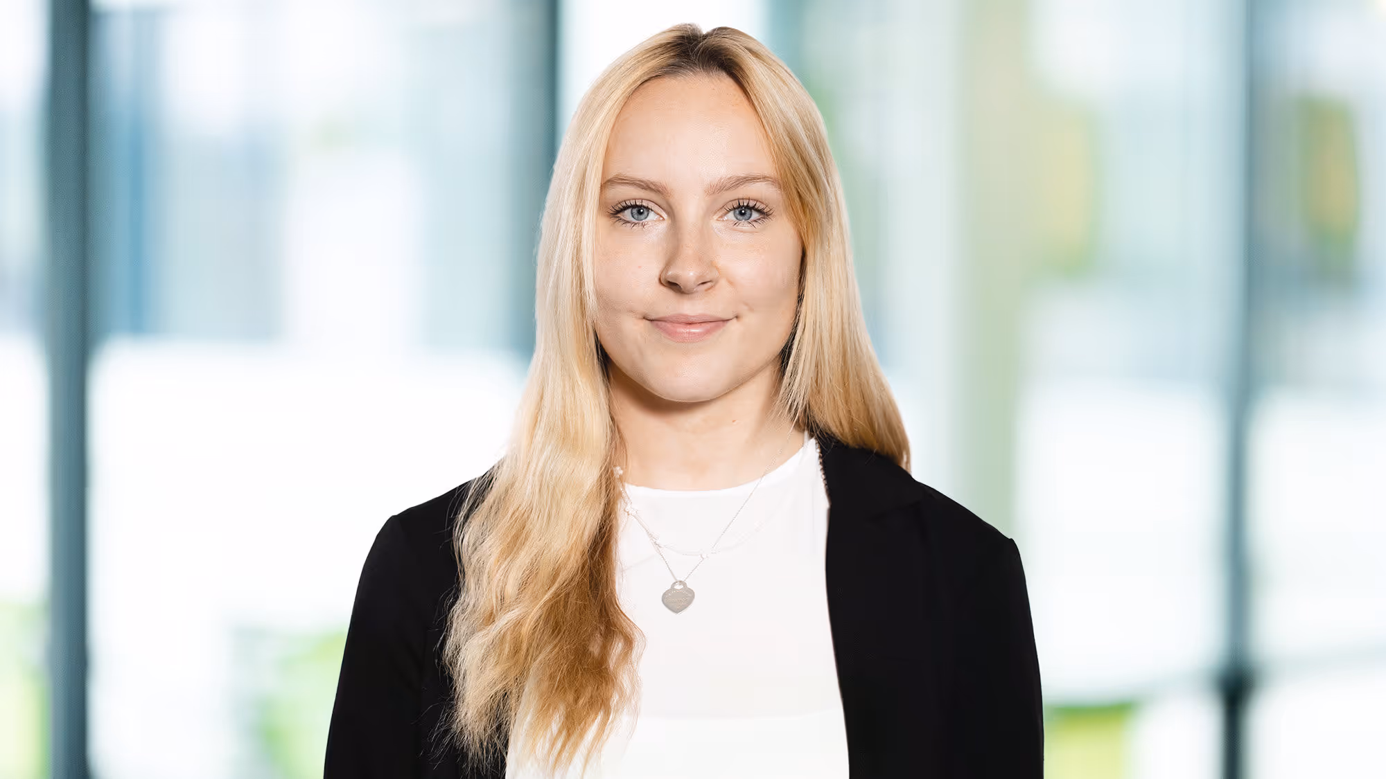 Junge Frau mit langen blonden Haaren, die einen schwarzen Blazer und ein weißes Oberteil vor einem verschwommenen Fensterhintergrund trägt.