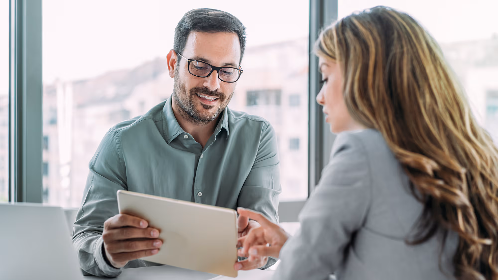 Man with glasses showing a tablet to a woman with long hair in a bright office setting.