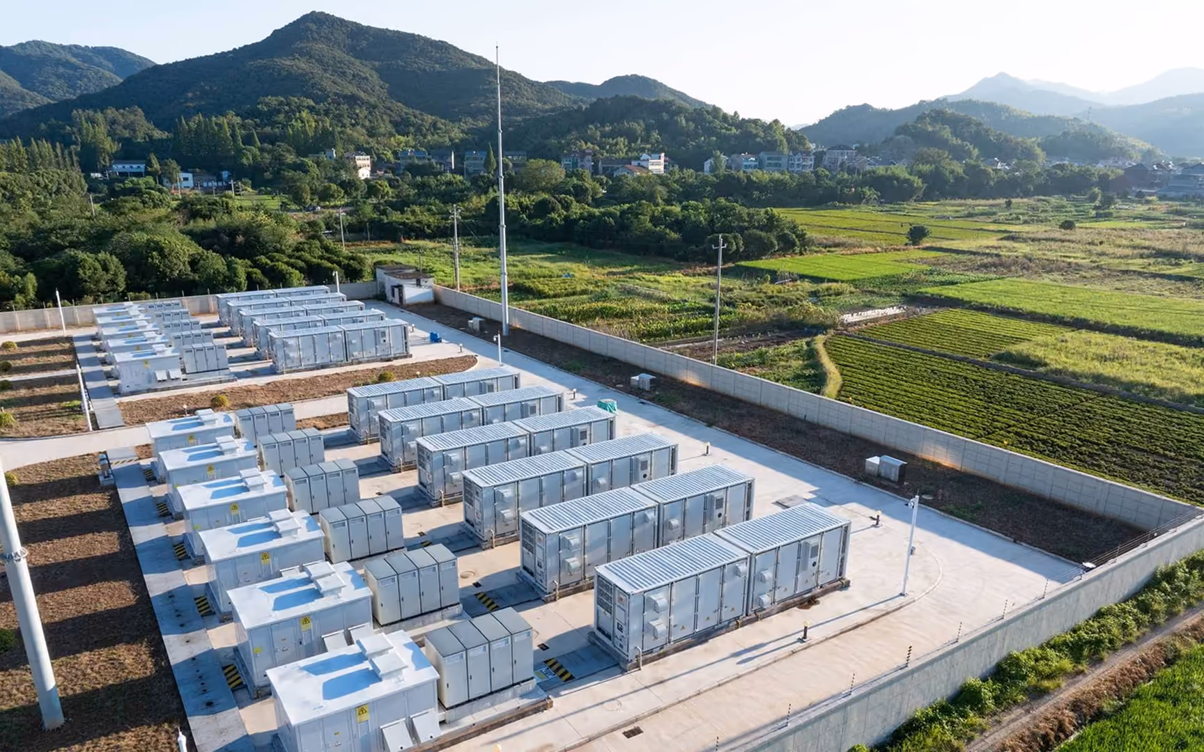 Energiespeicheranlage im Freien mit Reihen großer industrieller Batteriecontainer, umgeben von grünen Feldern und Bergen.