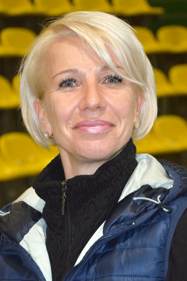 Smiling woman with short blonde hair wearing a blue jacket and black turtleneck with yellow stadium seats in the background.